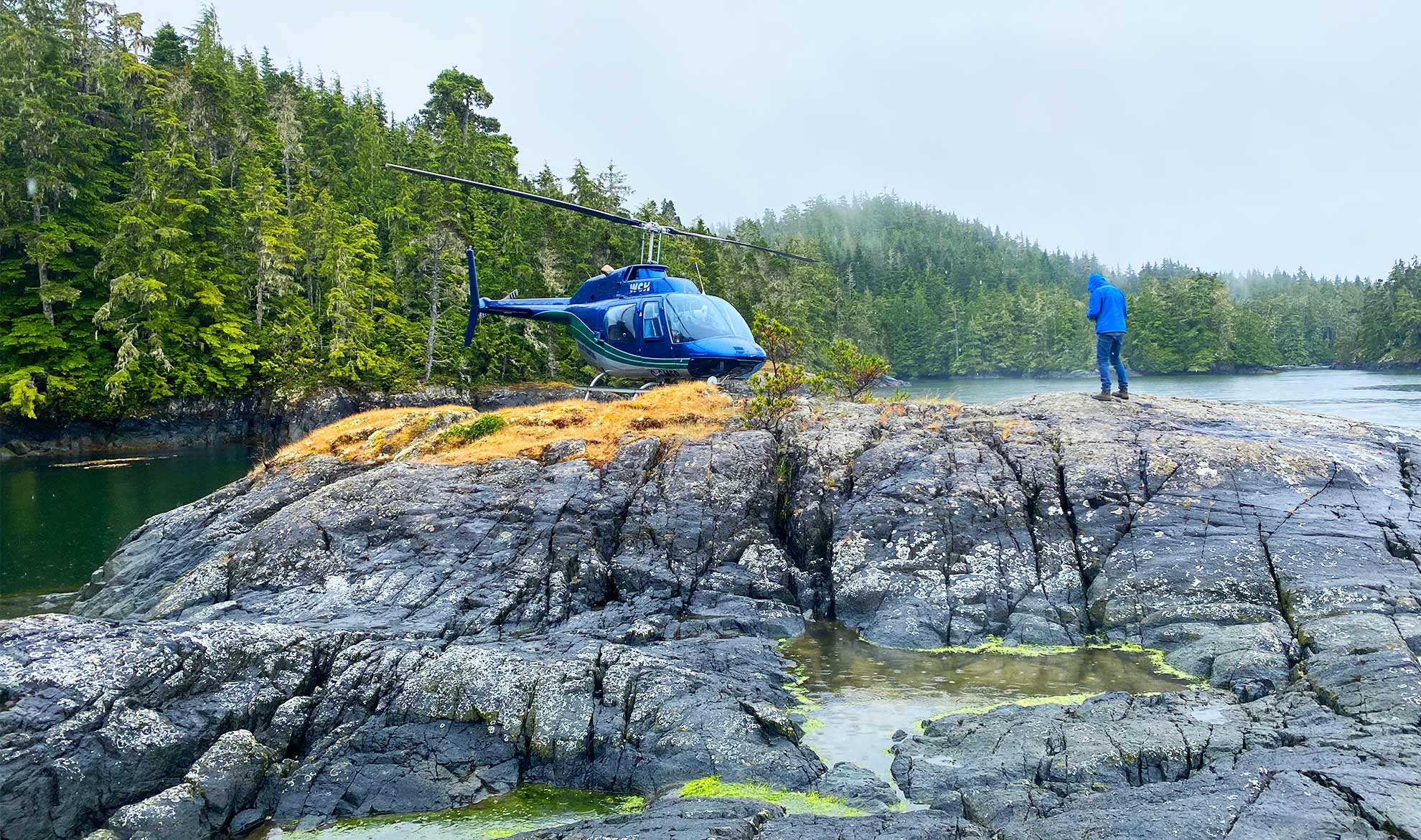 A blue helicopter landed on a rocky, moss-covered shoreline with a geoscientist in high-visibility gear standing near the coastal waters of British Columbia.